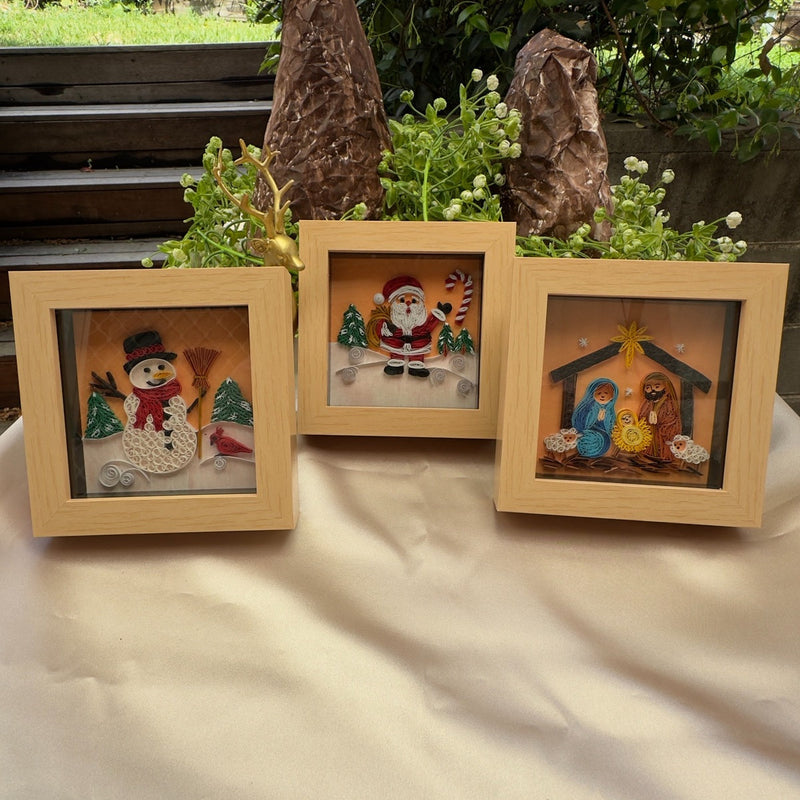 Three miniature paper quilling artworks featuring a snowman, Santa and a nativity scene in matching light wood-look frames, arranged on a table.