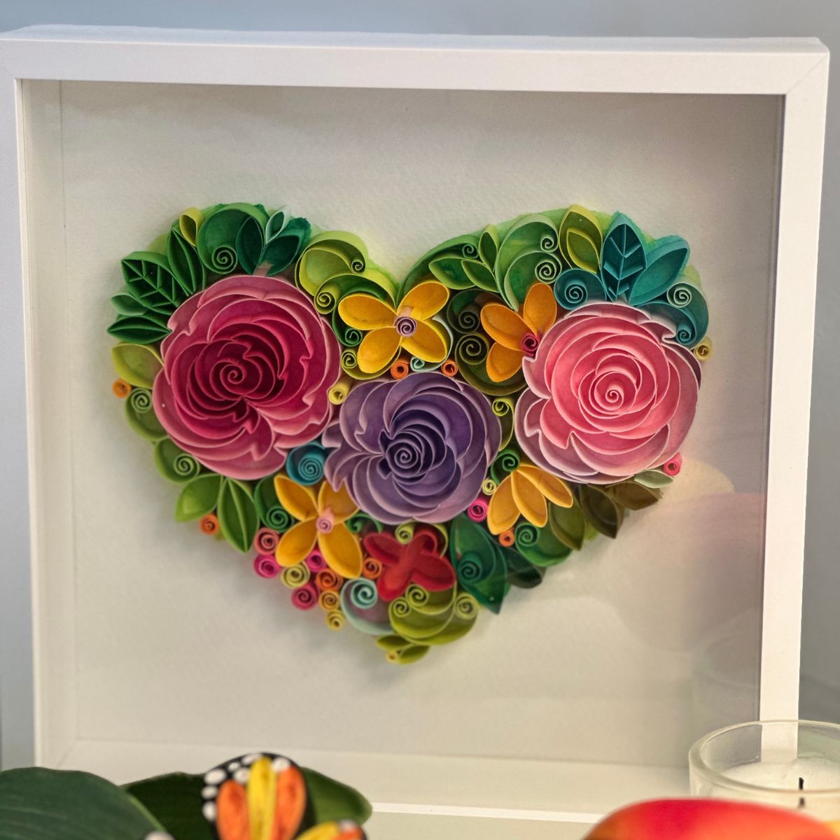 Handcrafted paper quilling artwork featuring a floral heart design with layered roses, flowers, leaves, and botanical details in a white shadow box frame.