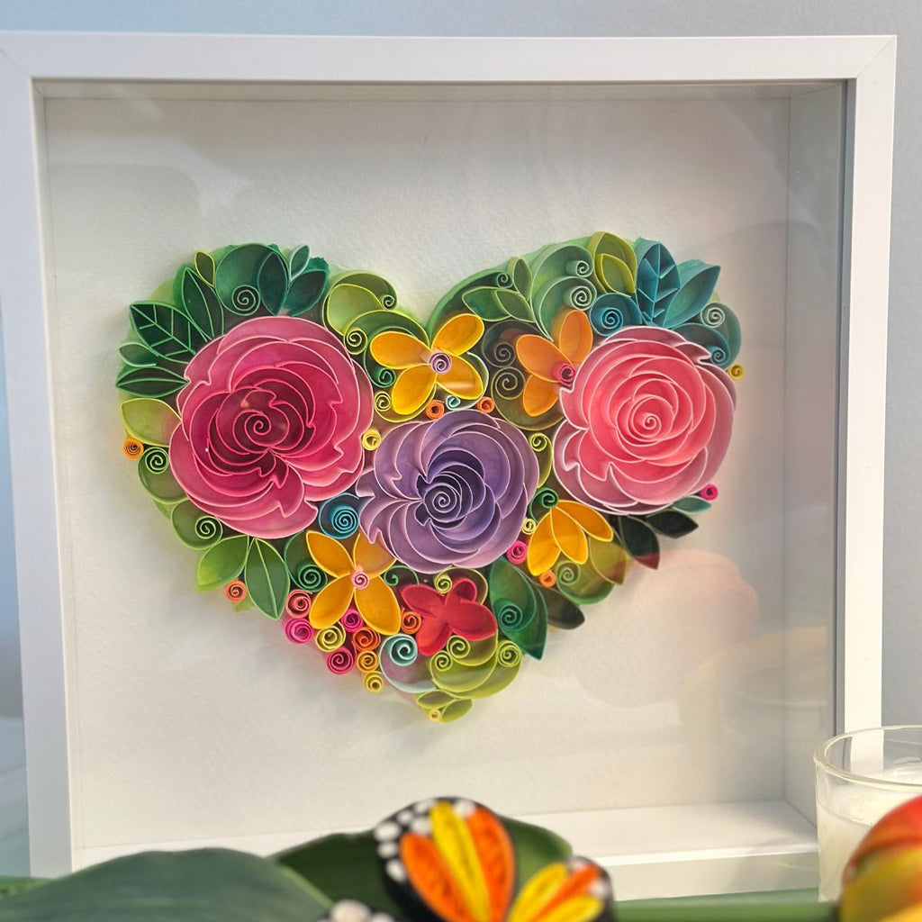 Handcrafted paper quilling artwork featuring a floral heart design with layered roses, flowers, leaves, and botanical details in a white shadow box frame.