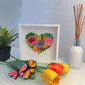 Handcrafted paper quilling artwork featuring a floral heart design with layered roses, flowers, leaves, and botanical details in a white shadow box frame.