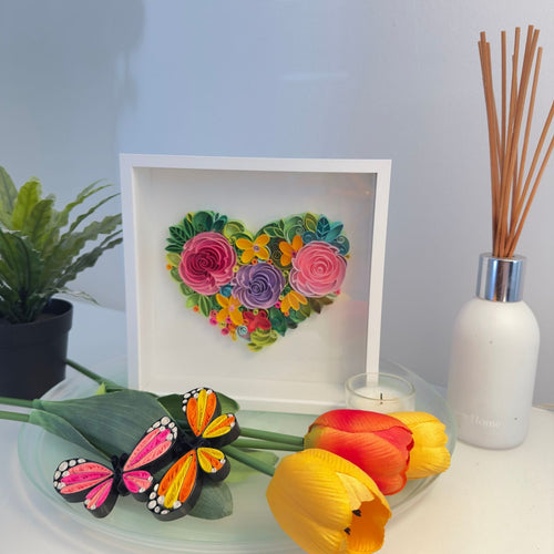 Handcrafted paper quilling artwork featuring a floral heart design with layered roses, flowers, leaves, and botanical details in a white shadow box frame.