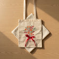 Hand embroidered floral tote bag on natural cotton canvas featuring bouquet-inspired orange and red floral stitching with red ribbon detail.