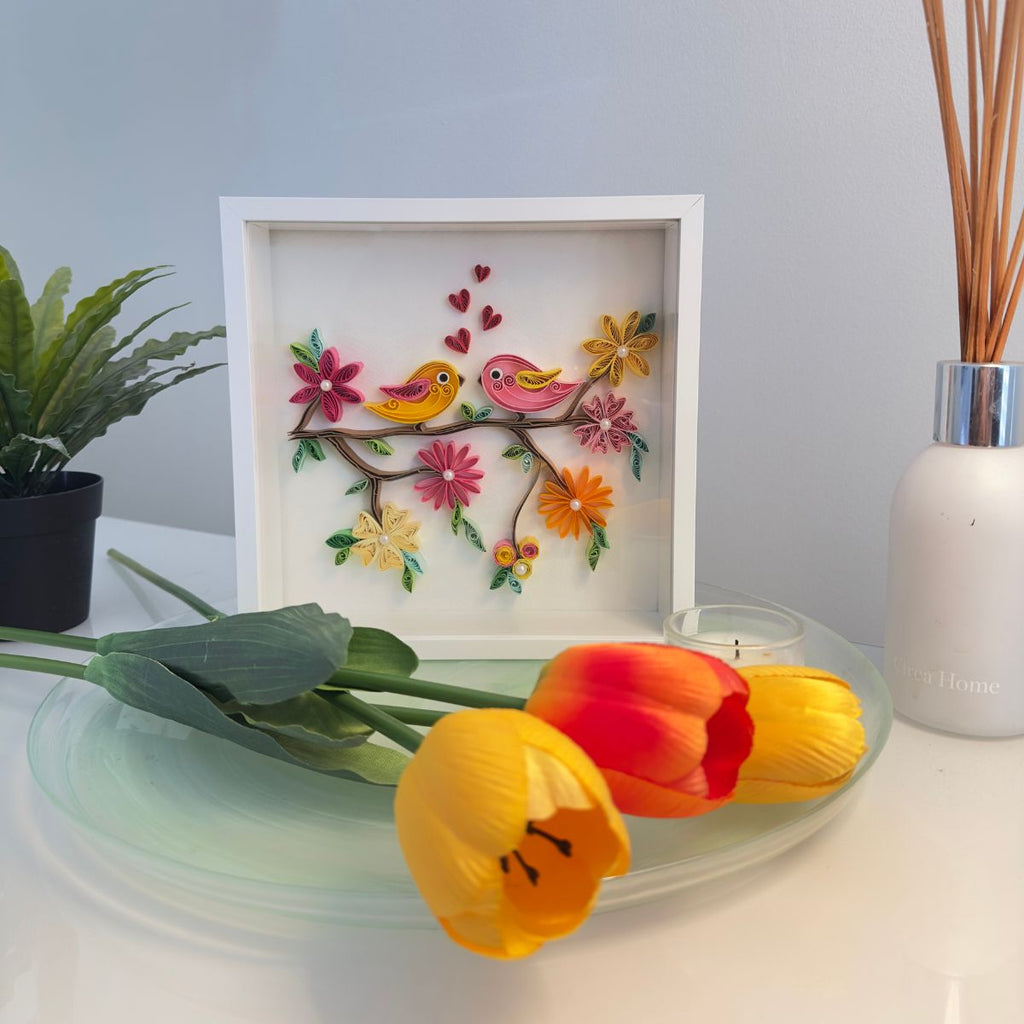 Decorative wall art with birds and flowers on a branch, placed on a surface with tulips and a plant.