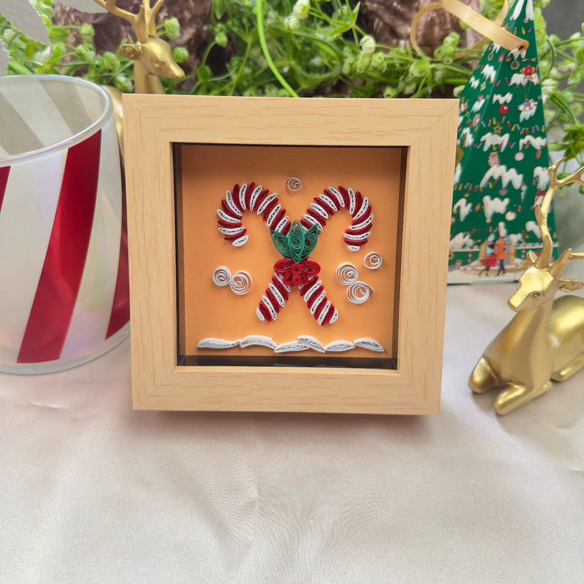 Red and white crossed candy cane paper quilling art with green leaves and red berries in a light wood-look frame, displayed among Christmas decorations.