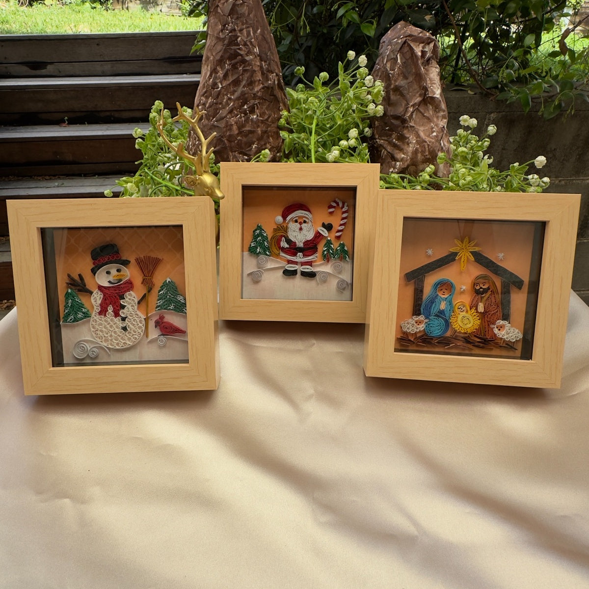 Three miniature paper quilling artworks featuring a snowman, Santa and a nativity scene in matching light wood-look frames, arranged on a table.