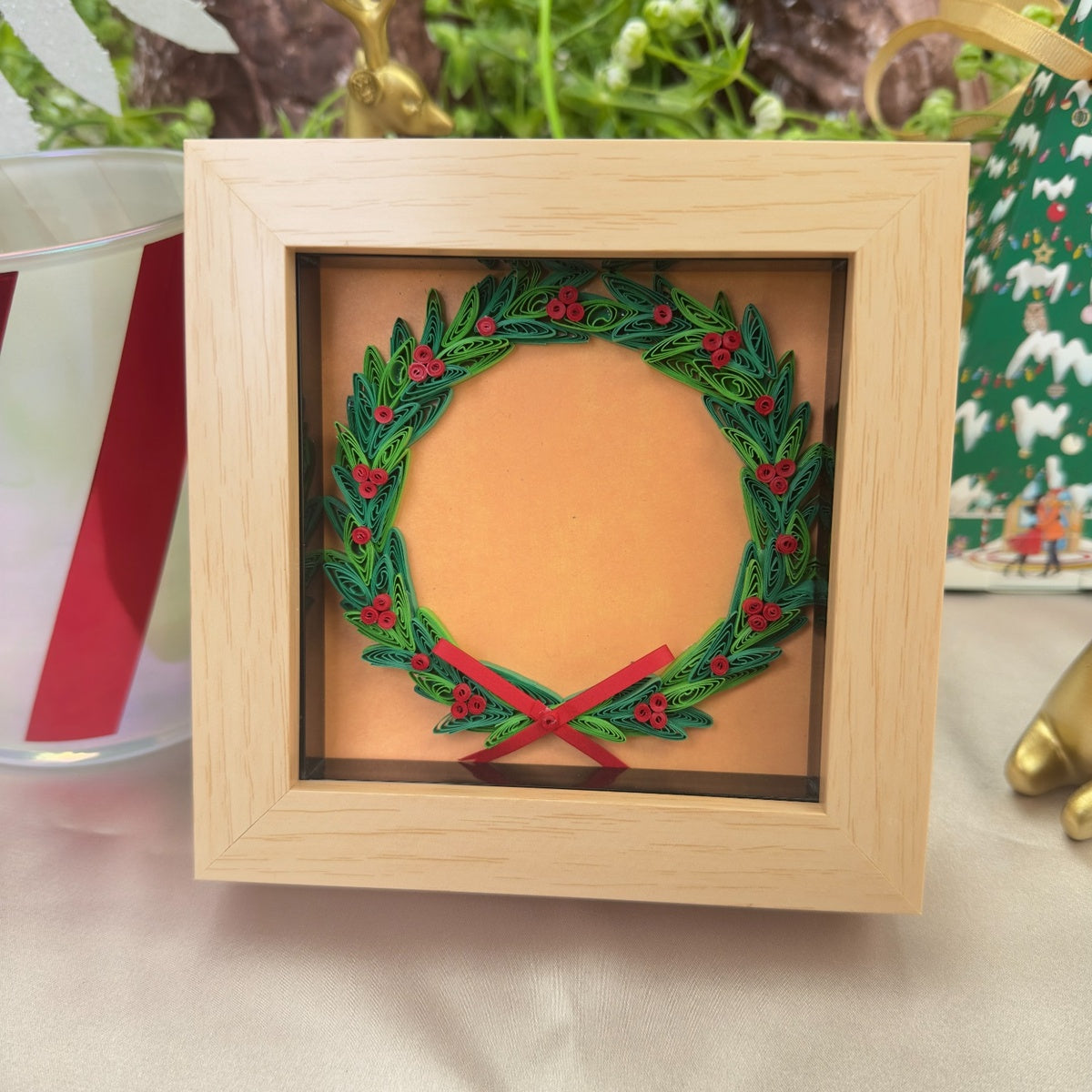 Christmas wreath paper quilling art with layered green leaves, red berries and a red bow in a light wood-look frame among festive decorations.