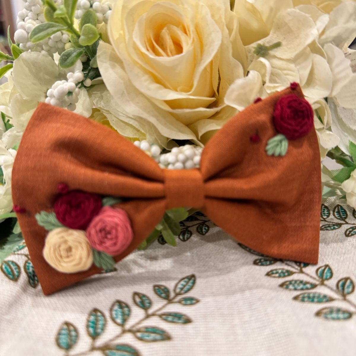 A rust-orange fabric hair bow with embroidered roses in burgundy, pink, and cream, accented with small green leaves