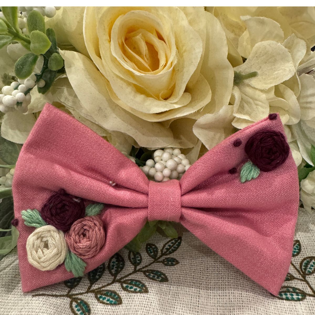 A pink fabric  hair bow with embroidered flowers in burgundy, cream, and dusty pink, with green leaves,