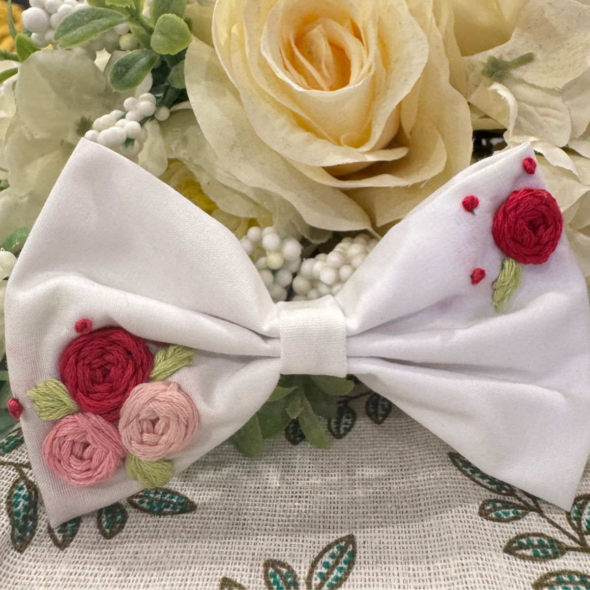 A white fabric hair bow featuring embroidered red and pink roses with green leaves
