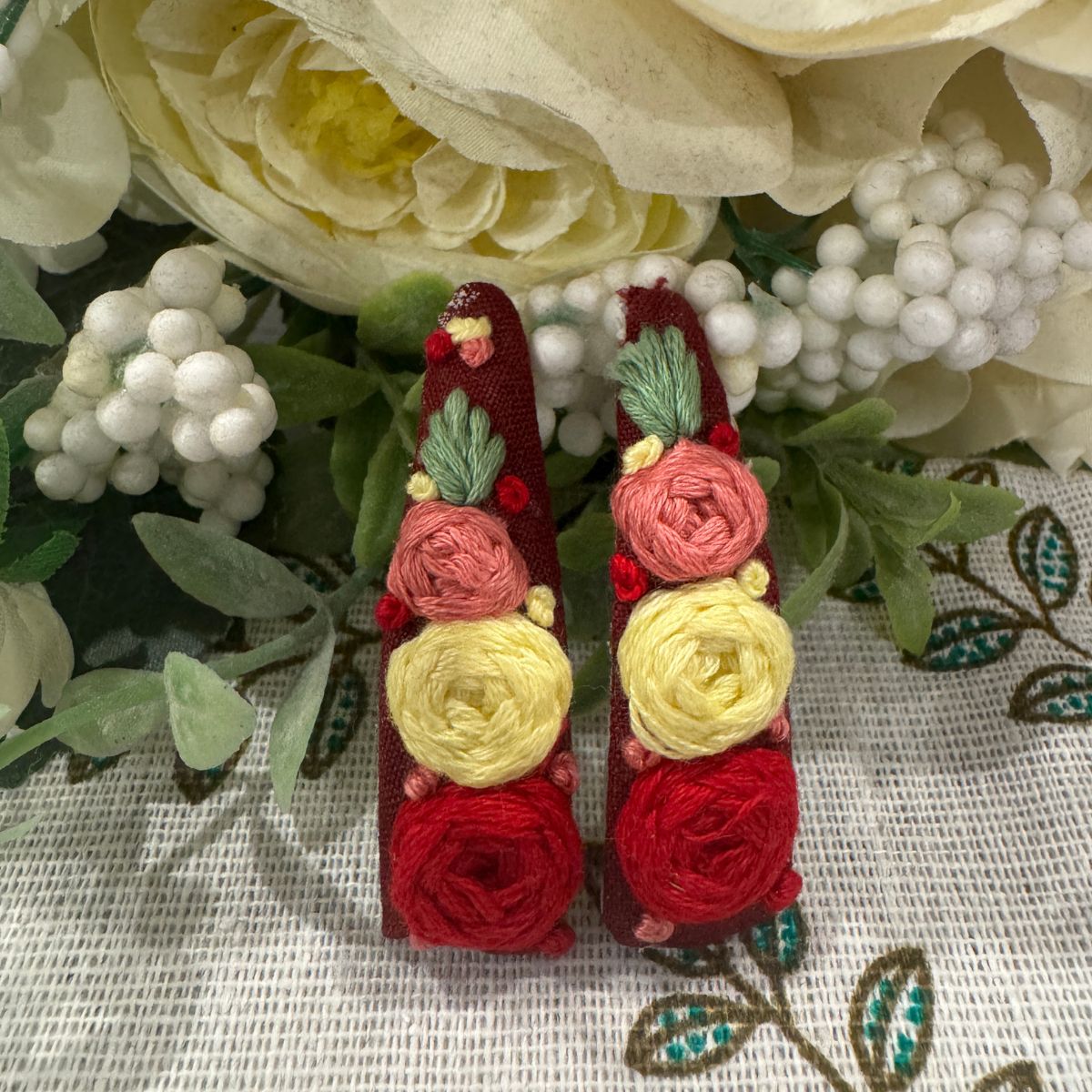 A pair of handmade red fabric hair clips decorated with rolled red, pink, and yellow fabric flowers and green leaves