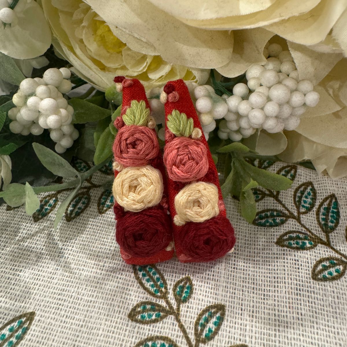 A pair of red fabric-covered tic tac clips with embroidered floral details in pink, cream, and green