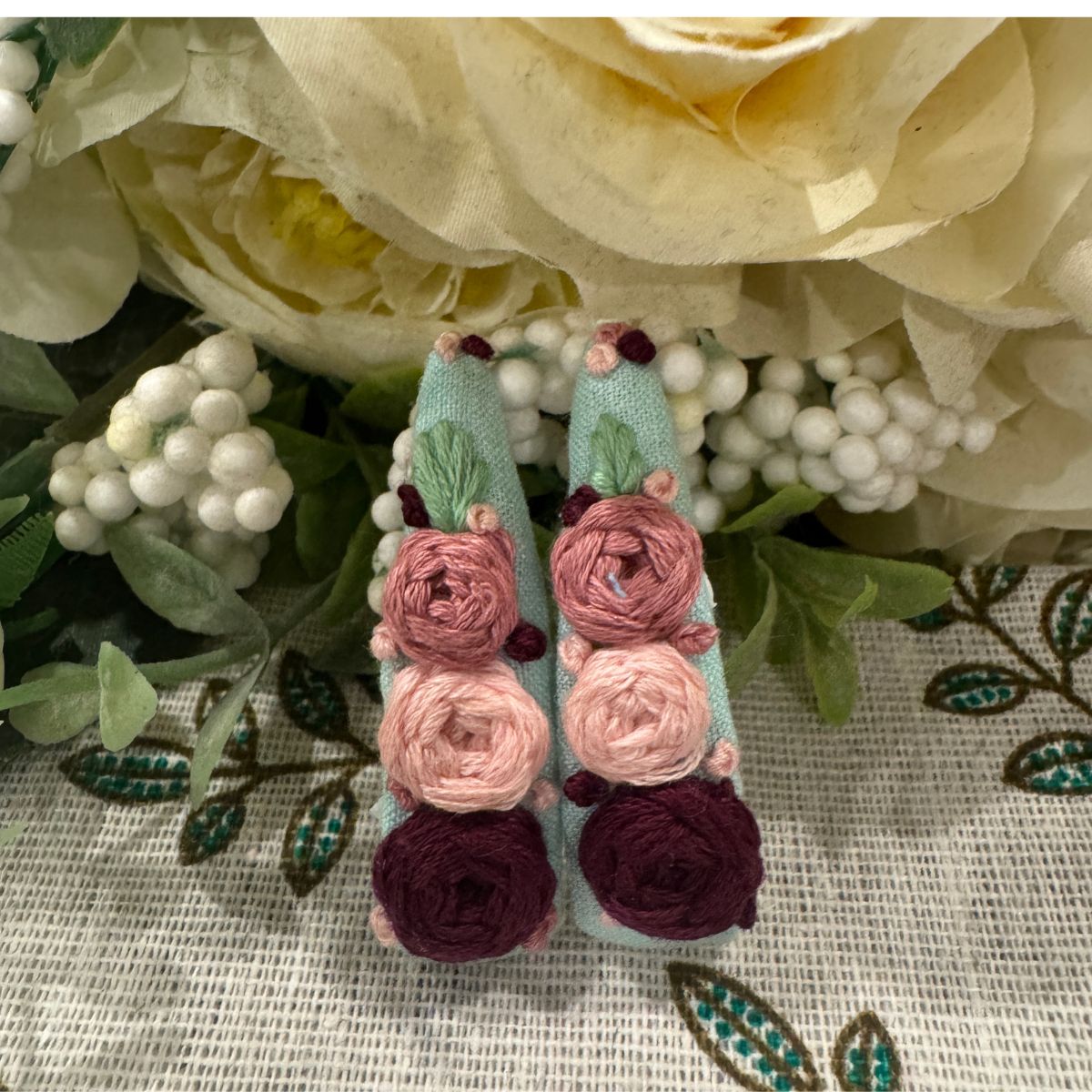 A pair of handmade mint green fabric hair clips with pink, dusty rose, and burgundy embroidered flowers