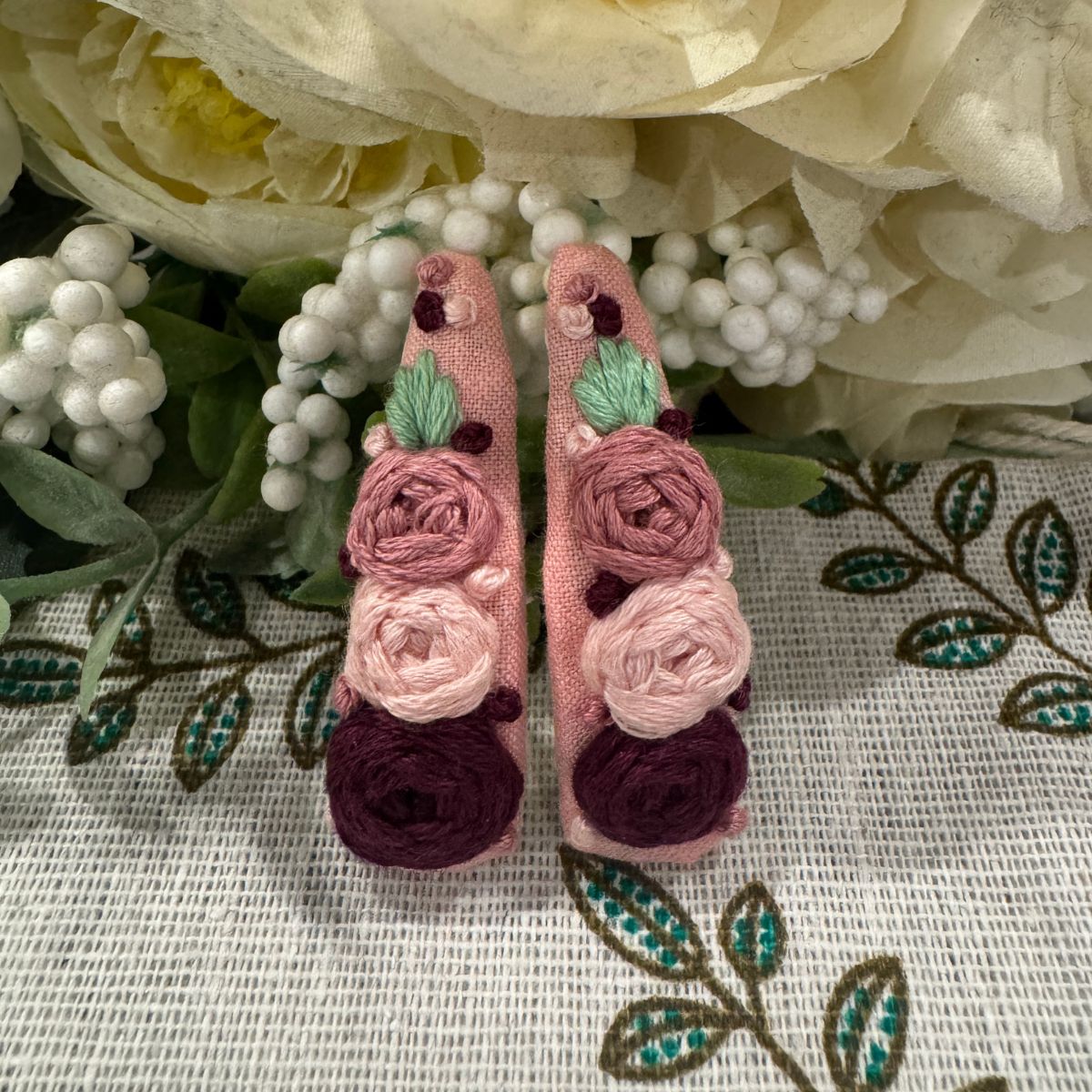 A pair of pink fabric hair clips with embroidered roses in shades of dusty pink, light blush, and deep burgundy, accented with small green leaves