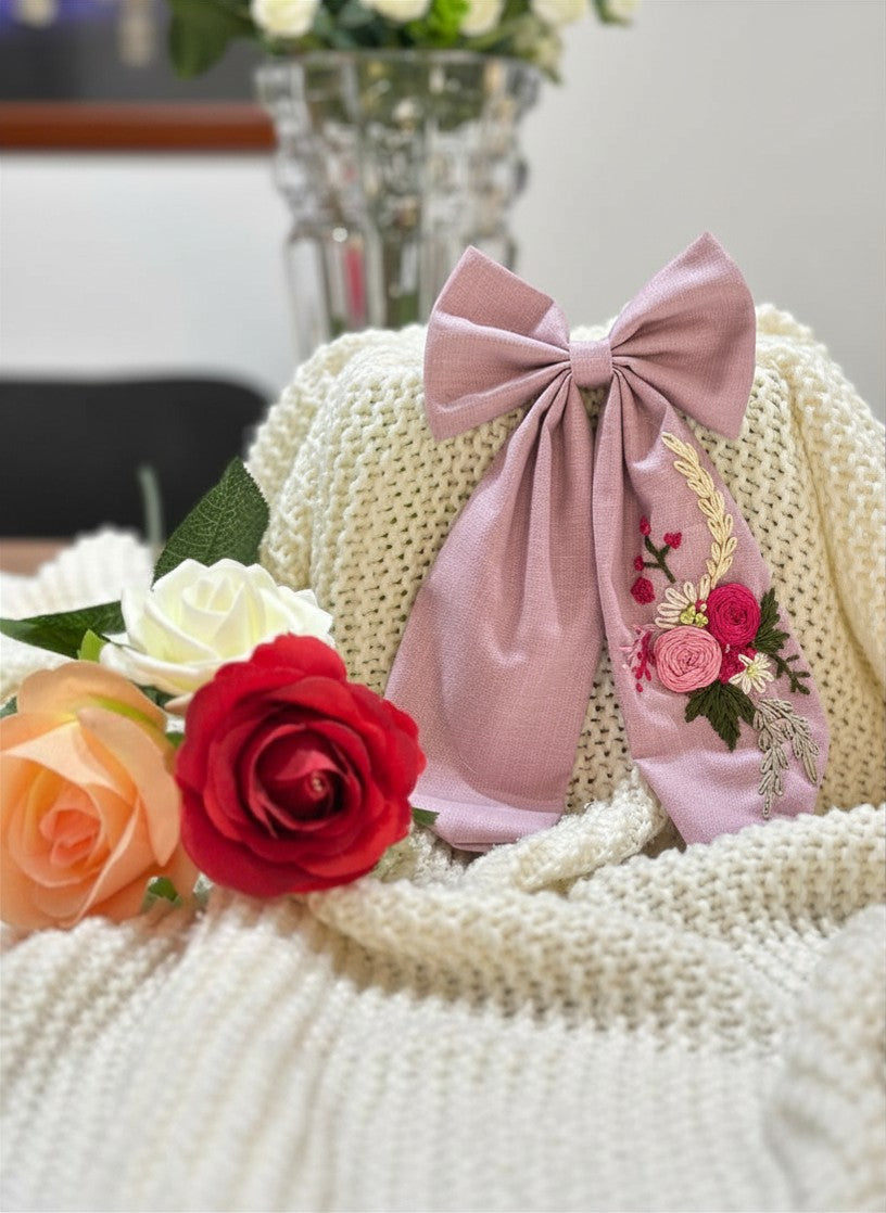 Hand embroidered floral pigtail bow in soft  pink cotton fabric featuring artisan floral stitching and alligator clip attachment for girls' hairstyles.
