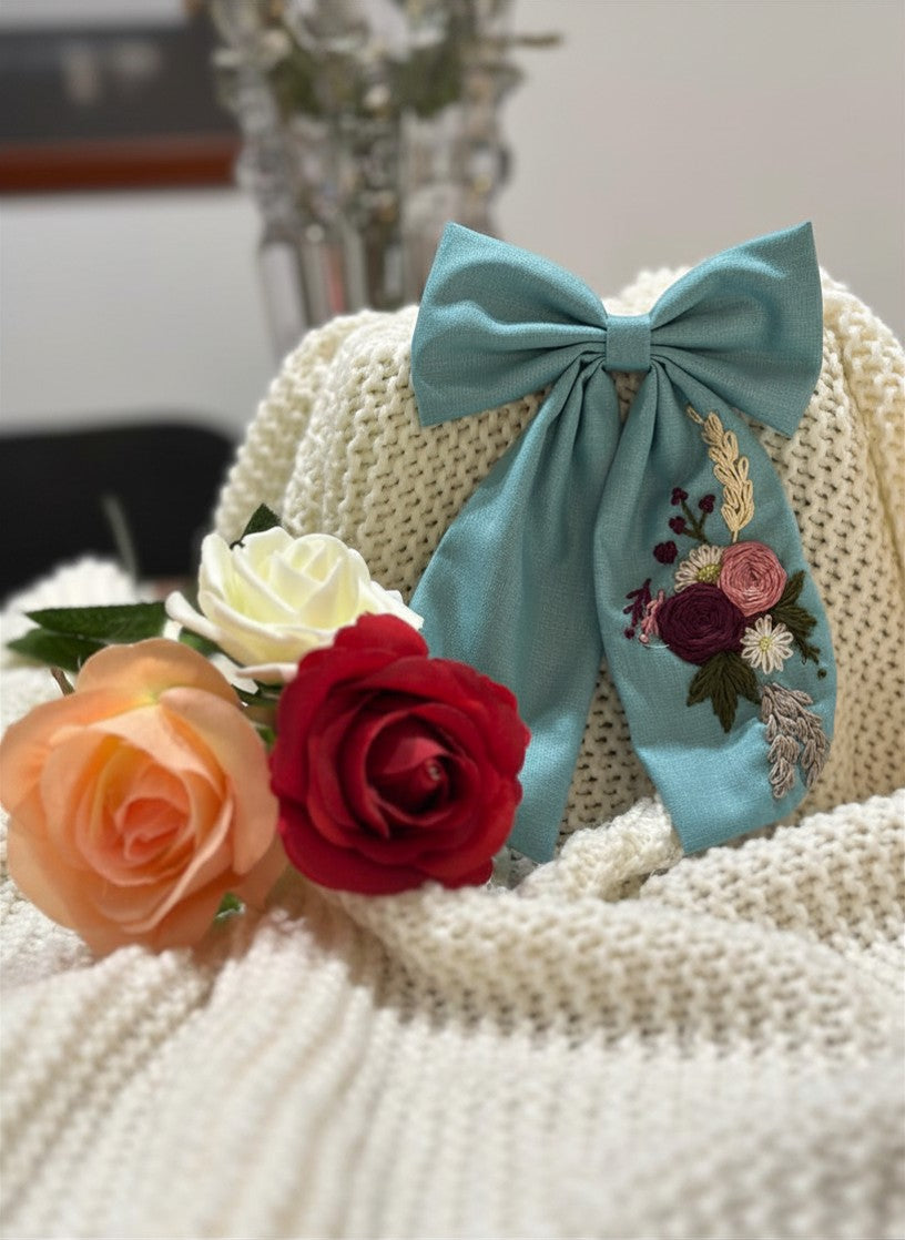 Hand embroidered floral pigtail bow in sky blue cotton fabric featuring artisan floral stitching and alligator clip attachment for girls' hairstyles.