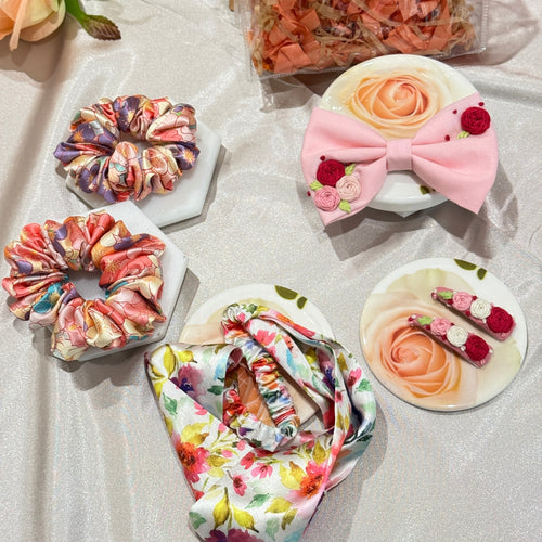 A styled flat-lay of hair accessories on a light satin fabric background, including two fabric scrunchies, a large pink fabric bow with floral details, a matching floral fabric headband, and two small floral hair clips.