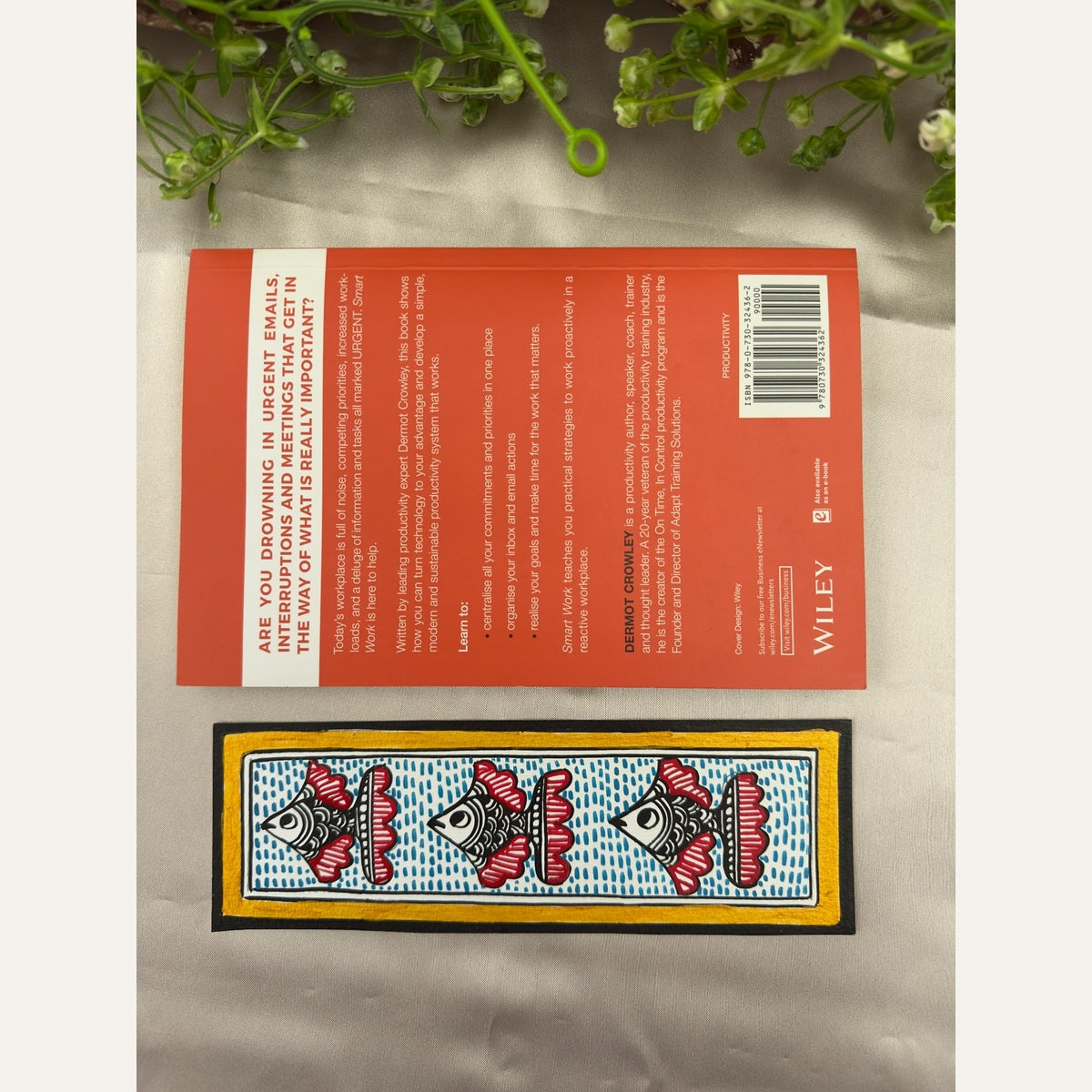Hand-painted Madhubani Scarlet Shoal bookmark with three black-and-white fish, crimson-red fins, blue dash detailing and a golden-yellow border, beside a book for size reference. Book not included.