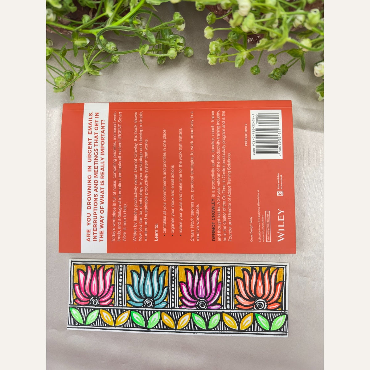Hand-painted Madhubani Fourfold Bloom bookmark with four pink, turquoise, magenta and coral lotus flowers, golden-yellow panels and a green-and-yellow leaf border, beside a book for size reference. Book not included.