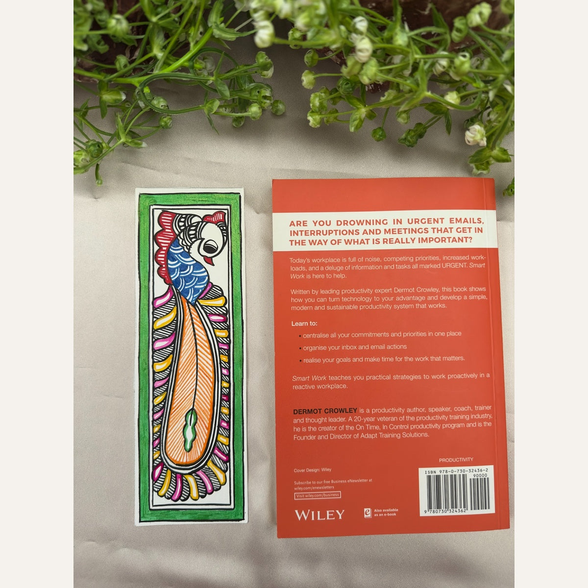 Hand-painted Madhubani Amber Plume peacock bookmark with a blue body, long orange tail, pink and yellow feather accents and a green border, beside a book for size reference. Book not included.