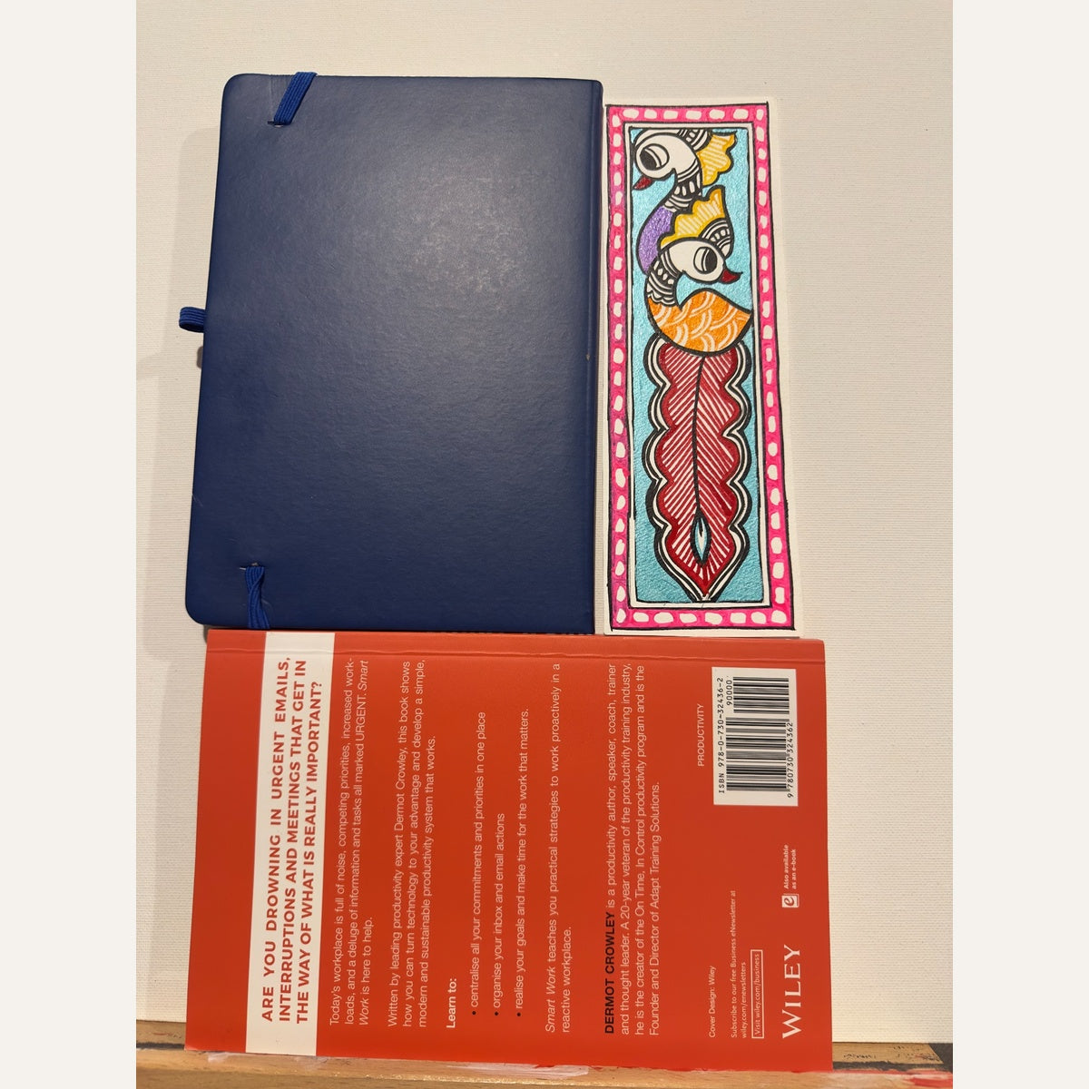 Hand-painted Madhubani peacock bookmark with purple and orange birds, a long crimson-red plume, turquoise background and pink dotted border, beside a notebook and book for size reference. Book and notebook not included.