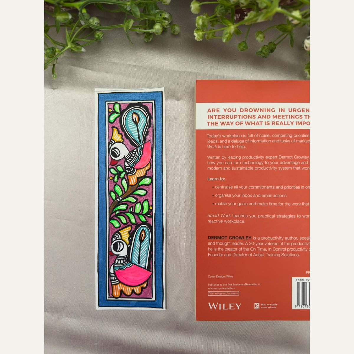 Hand-painted Madhubani Peacock Garden bookmark with two pink peacocks, turquoise tail feathers and green leaves, on a rose-purple background with a deep blue border, beside a book for size reference. Book not included.