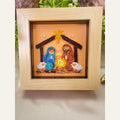 Framed nativity paper quilling art depicting Mary in blue, Joseph in terracotta, baby Jesus, two white sheep and a golden star above a brown stable.