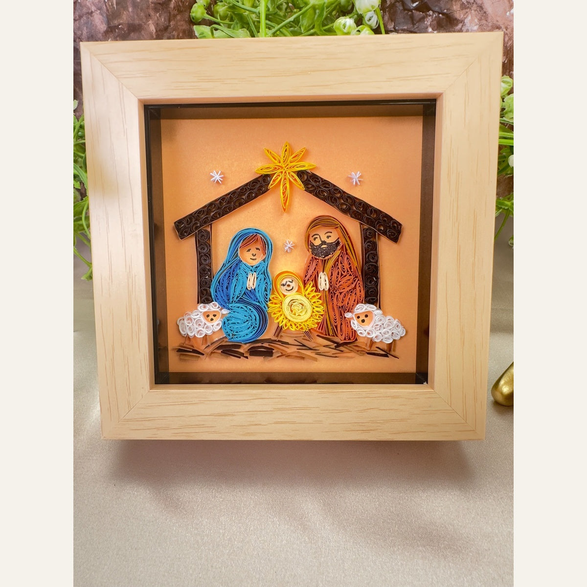Framed nativity paper quilling art depicting Mary in blue, Joseph in terracotta, baby Jesus, two white sheep and a golden star above a brown stable.