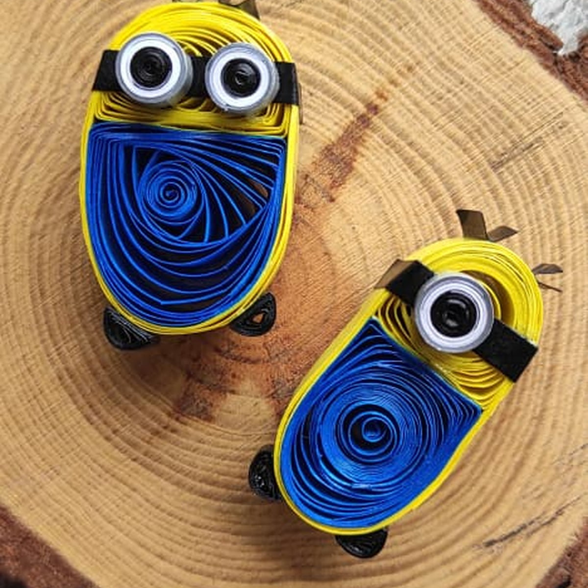 One-eye and two-eye handcrafted paper quilling character fridge magnets displayed together on a natural wood slice.