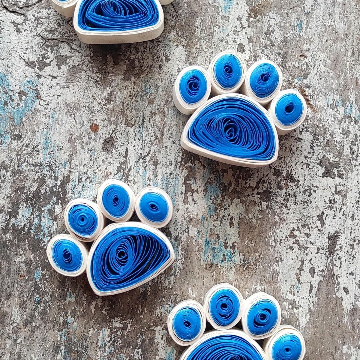 Handmade blue and white paper quilling paw fridge magnets displayed on a textured rustic background.