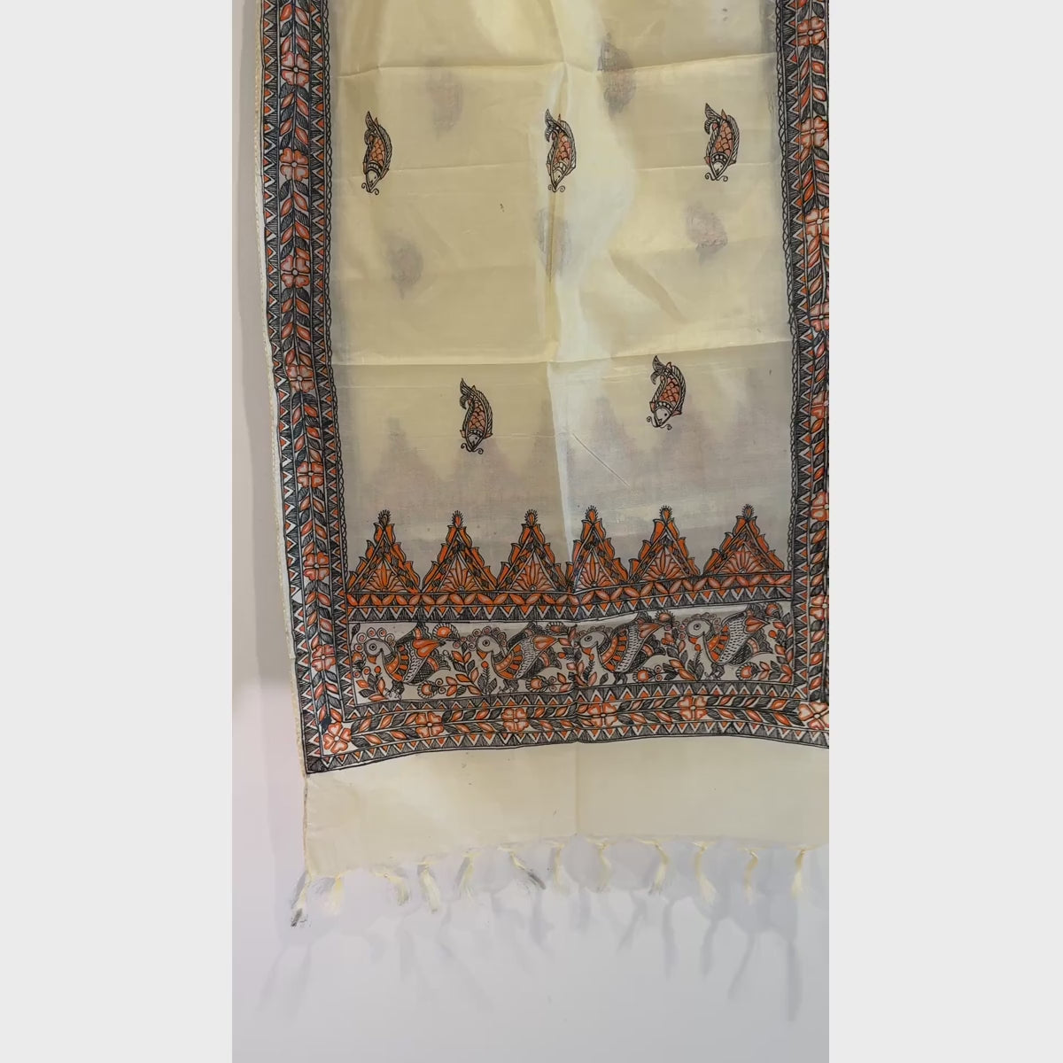 Close-up video of an ivory cream hand-painted Madhubani art silk stole featuring orange and black fish motifs, botanical details, geometric borders and tasselled ends.