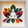 A framed wall art piece featuring a lotus-shaped silhouette with a seated meditating figure in the center, made from layered rainbow-colored strips. The artwork is displayed in a white square frame against a warm off-white background.