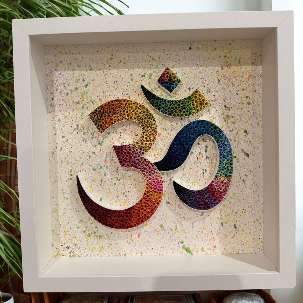 A framed Om symbol wall art piece featuring a multicolor rainbow gradient quillwork design on a speckled cream background, housed in a white square frame.