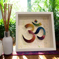 Handcrafted rainbow Om paper quilling artwork in a white shadow box frame featuring intricate paper coils on a hand-painted splatter background, ideal for meditation and spiritual home décor.