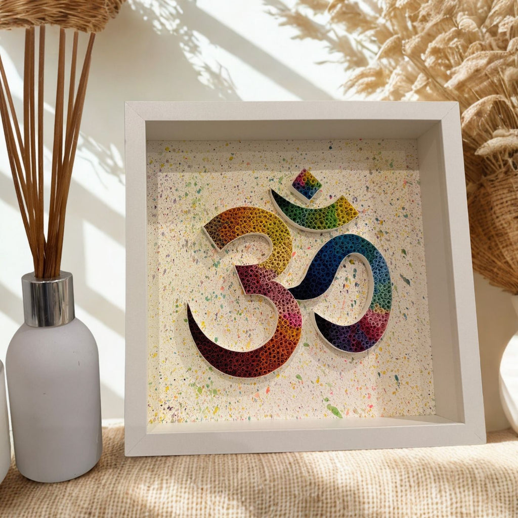 A square white shadow box art piece featuring a colorful, textured Om symbol on a speckled cream background, styled on a burlap surface next to a white reed diffuser.