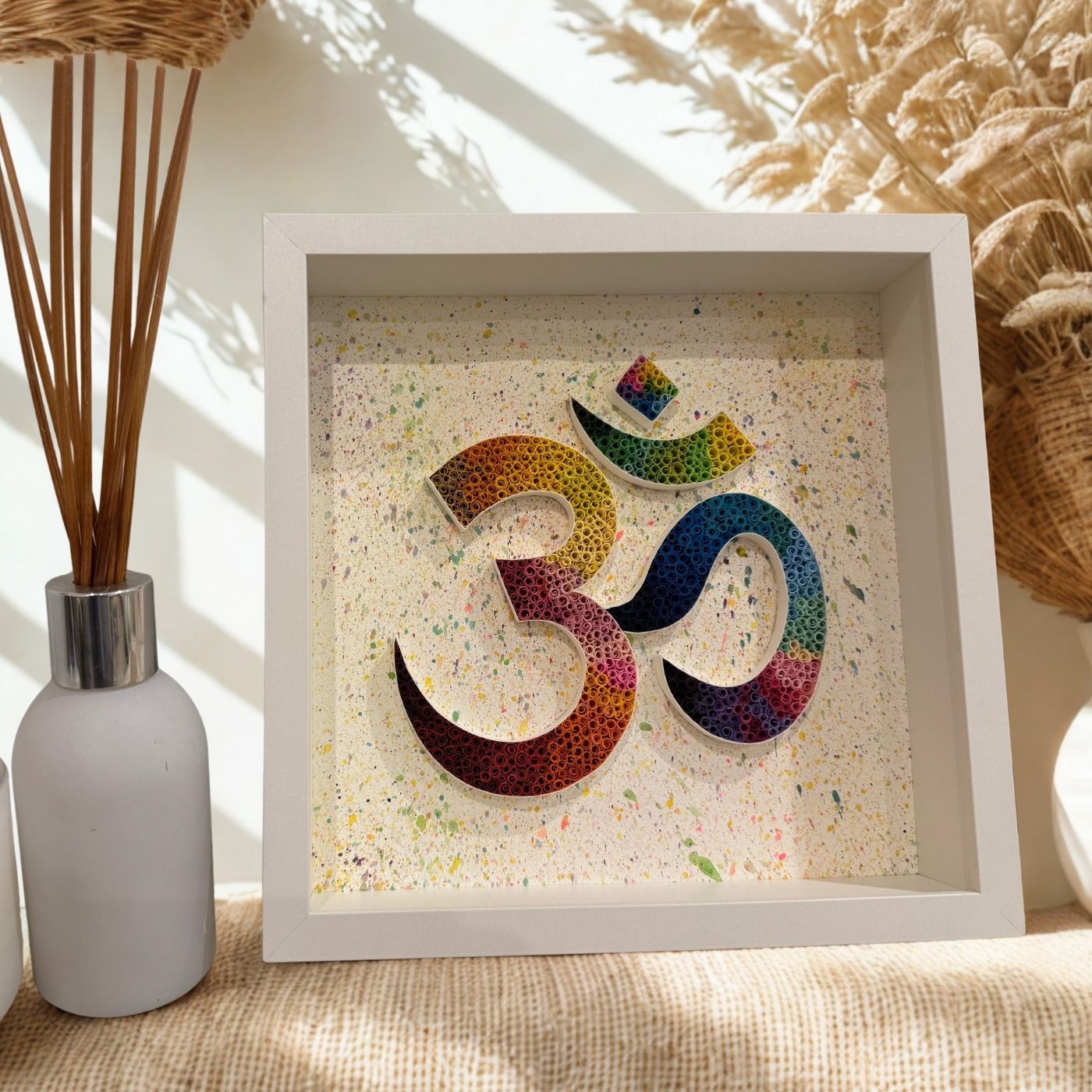 A square white shadow box art piece featuring a colorful, textured Om symbol on a speckled cream background, styled on a burlap surface next to a white reed diffuser.