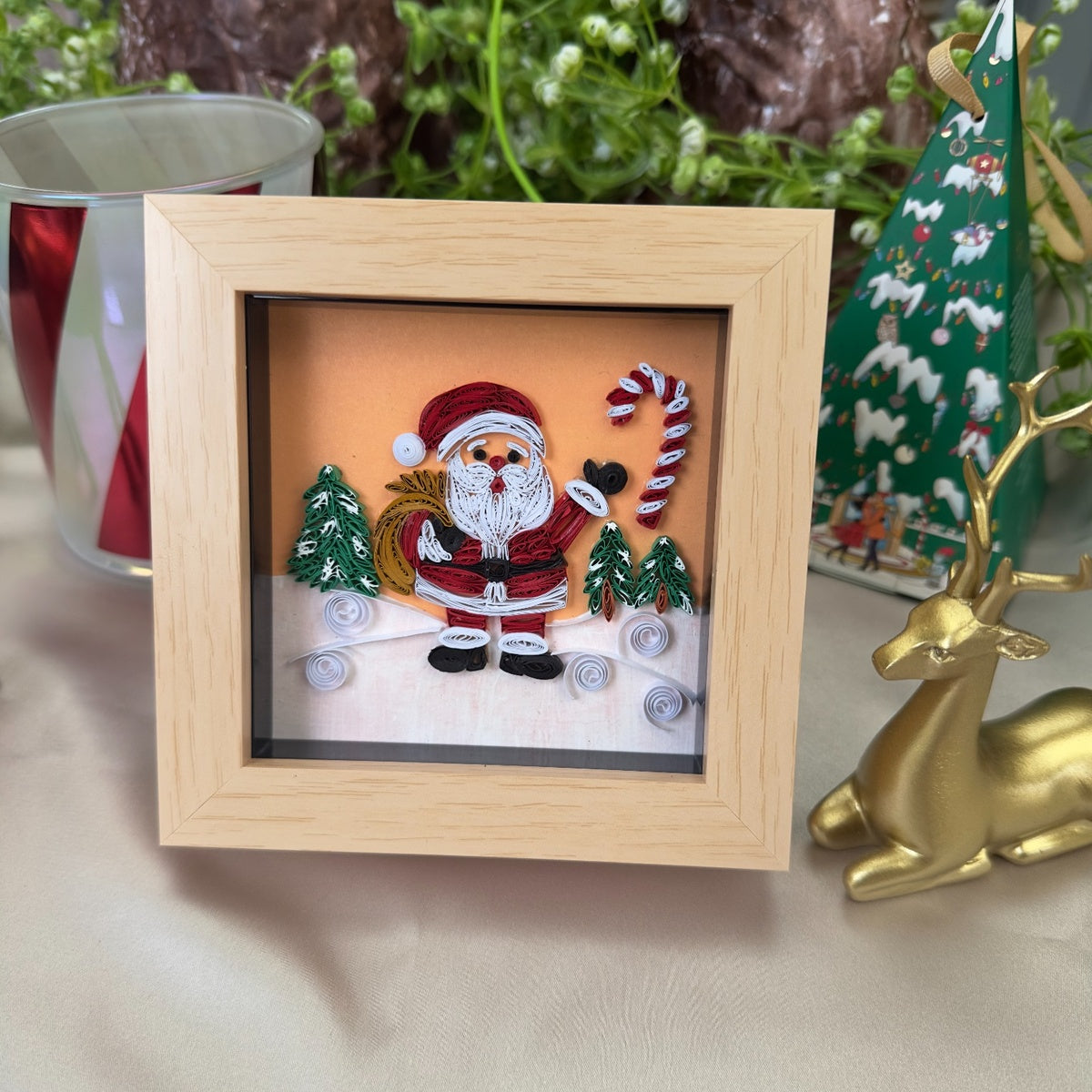Handcrafted Santa paper quilling art with a red suit, white beard, golden yellow sack, green trees and candy cane in a light wood-look frame.
