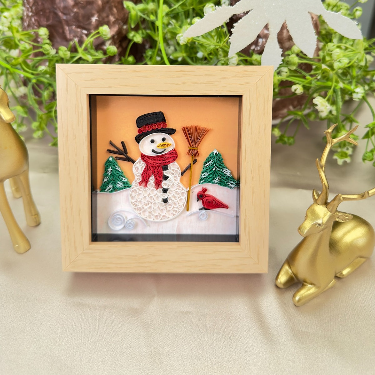 Mini snowman paper quilling art with a red scarf, green trees and a red bird in a light wood-look frame, displayed among Christmas decorations.